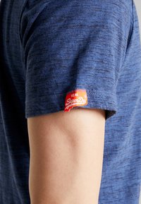 Niebieska koszulka z krótkim rękawem z teksturowanej tkaniny. Charakterystyczna czerwona i biała metka "SuperDry" na rękawie dodaje szczegółu.