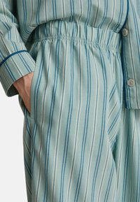 Hand in de zak, gekleed in lichtblauwe gestreepte pyjamabroek met elastische tailleband en bijpassend langarmtop met knopen en marineblauwe afwerking.