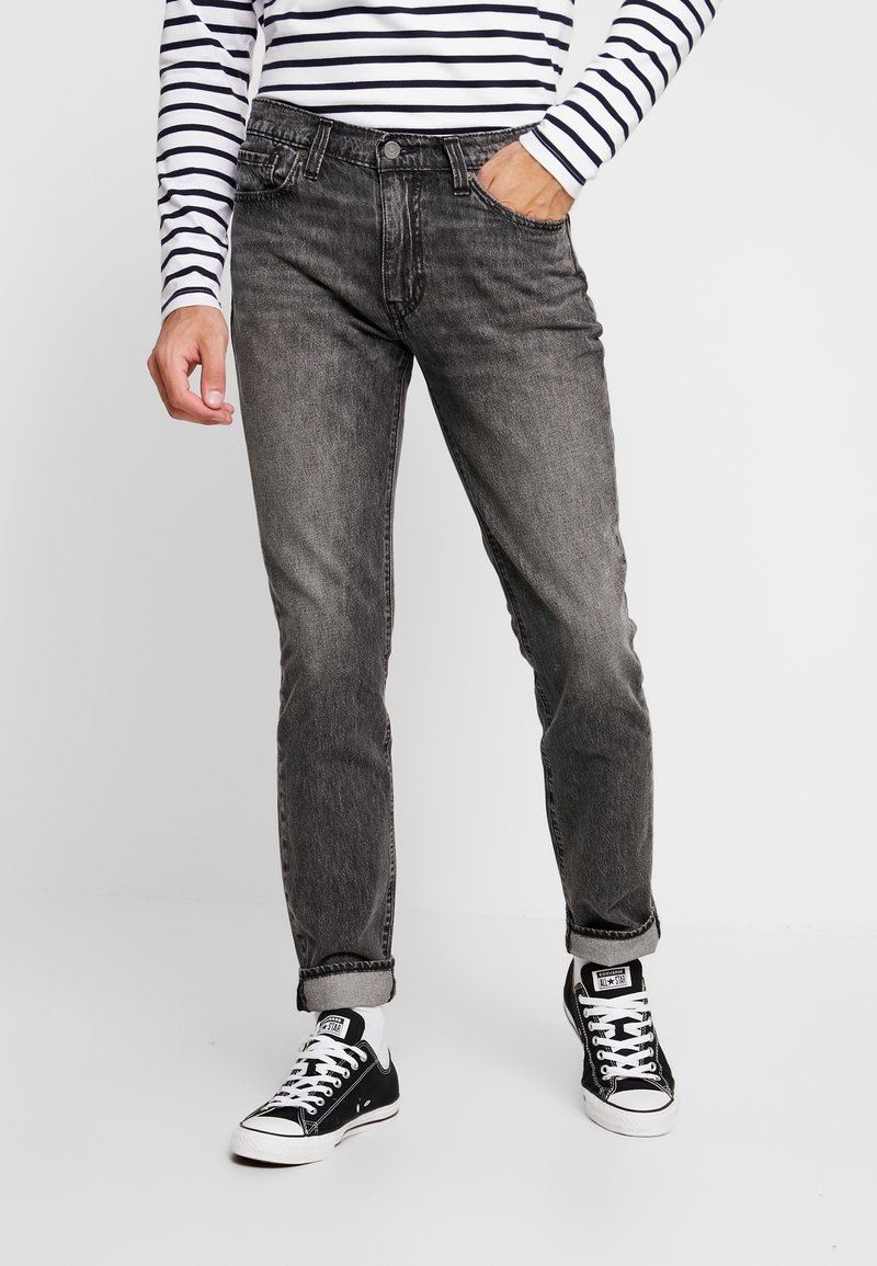 Jeans en denim gris à coupe droite, présentant une texture légèrement délavée et des ourlets retroussés. Associés à des baskets en toile noires.