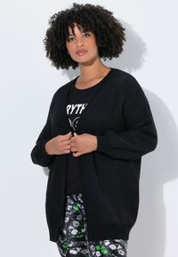 Cardigan noir à coupe ample et manches longues, fabriqué dans un tissu texturé. Porté sur un t-shirt noir avec motif et un pantalon à motifs.