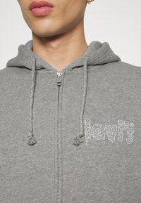 Sweat à capuche gris avec fermeture éclair, fabriqué en tissu doux, doté d'une capuche à cordon de serrage et d'un logo blanc texturé sur la poitrine. La fermeture éclair est métallique.