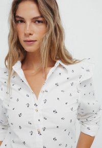 Witte langemouwen blouse met kleine bloemenpatronen in zwart en paars, voorzien van knoopsluiting en gerimpelde mouwen voor een accent.