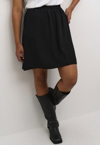 Jupe noire, arrivant au genou, avec une taille élastique, fabriquée dans un tissu lisse. Portée avec des bottes en cuir noires, montant jusqu'au genou.