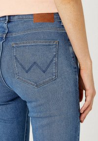 Blå denimjeans med en närbild av en bakficka som har ett zigzagformat "W"-stickningsmönster. Inkluderar en brun läderlogotyp på midjebandet.