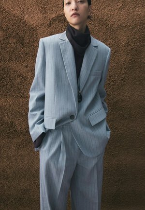 Personne portant un costume oversize bleu clair à fines rayures blanches et un col roulé noir, debout contre un mur brun texturé, les mains dans les poches.
