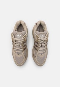 Ljusa beige sportskor med mesh och syntetmaterial, med en texturerad tunga, snören och tre bruna accentstripes.