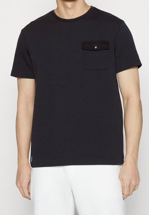 Homme portant un t-shirt noir à manches courtes avec une poche poitrine boutonnée et un pantalon blanc sur un fond uni.