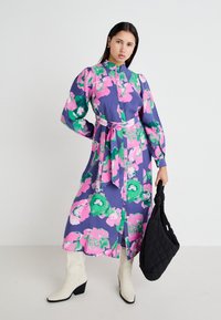 Blommig klänning med lila bas, stora rosa och gröna blommor, långa ärmar, hög krage och midja med bälte; i kombination med vita klackstövlar och en svart väska.