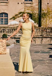 Femme en robe jaune sans manches debout à l'extérieur sur une terrasse en pierre, touchant un coussin à motif floral sur un banc beige avec un bâtiment en arrière-plan.