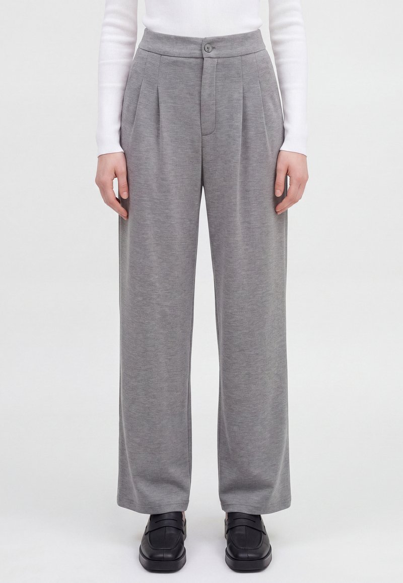 Pantalones grises de tiro alto y pierna ancha con pliegues delanteros, cierre de botón y cremallera, combinados con una camiseta blanca de manga larga y mocasines negros.