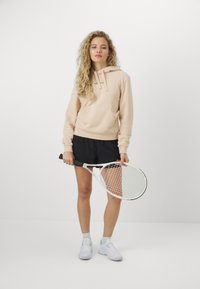 Felpa beige chiaro con tasca a marsupio, abbinata a pantaloni corti neri. Il modello tiene in mano una racchetta da tennis, indossando sneakers bianche con dettagli blu.