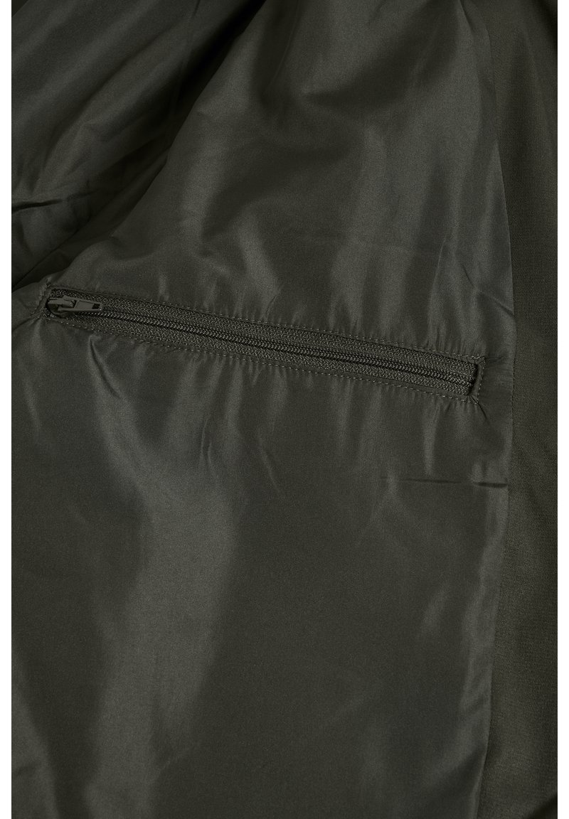 Tissu de veste vert foncé avec un détail de poche zippée, mettant en avant une texture lisse et une finition mate. Éléments de design simples et fonctionnels.