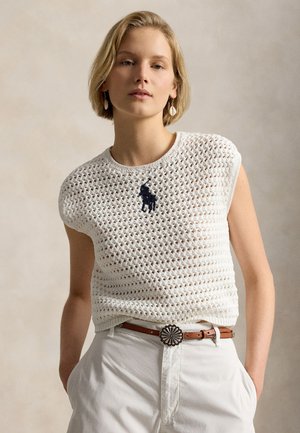 Femme aux cheveux courts blonds portant un haut sans manches en crochet blanc avec un logo polo noir, un pantalon blanc, une ceinture marron et des boucles d'oreilles en forme de coquillage.