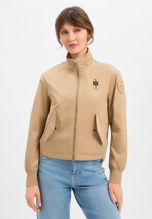 Vrouw draagt een beige jas met ritssluiting en klepzakken, gecombineerd met een blauwe spijkerbroek, staand tegen een witte achtergrond.