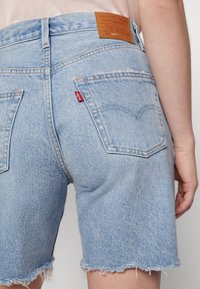 Light blue denim shorts with a frayed hem, featuring back pockets and a tan leather patch on the waistband with brand logo.