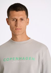 Sudadera gris con cuello redondo, con el texto "COPENHAGUE" en verde negrita y "LINDBERGH" en gris claro. Textura de tela suave.