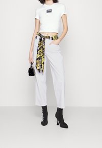 Mujer con un top corto blanco y pantalones claros, llevando un cinturón de seda estampado, sosteniendo un pequeño bolso negro y usando botas de tacón negro hasta el tobillo.