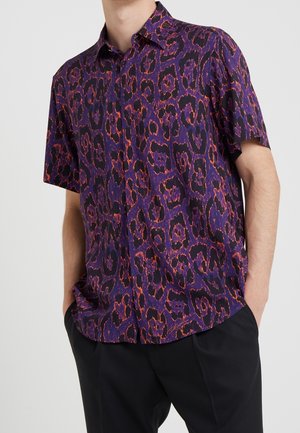 Mężczyzna w koszuli na guziki z krótkim rękawem z fioletowo-pomarańczowym, abstrakcyjnym nadrukiem w panterkę, w połączeniu z czarnymi spodniami, z rękami w kieszeniach.