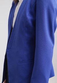 Blazer ajustado en tela azul royal, con un solo bolsillo frontal, diseño elegante y líneas de hombro estructuradas. Textura suave.