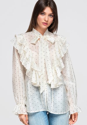 Kvinde iført en gennemsigtig hvid bluse med små sorte hjerter, store flæser, en sløjfe ved kraven og puffede ærmer, kombineret med lyseblå jeans.