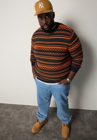 Pull en laine avec des motifs géométriques multicolores en orange, vert et noir ; associé à un jean bleu clair et des bottes beige. Casquette marron.