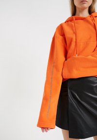 Sweatshirt à capuche orange avec une poche frontale et un cordon de serrage, accompagné de texte gris le long de la manche. Assorti à une jupe en faux cuir noire.