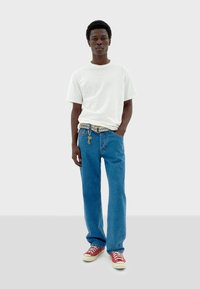 Camiseta blanca de manga corta, vaqueros azules de pierna recta, zapatillas rojas, con un cinturón decorativo que muestra un llavero. Texturas de tela suaves.