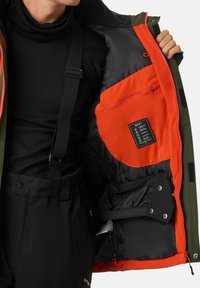 Grüne Außenjacke mit orangefarbenem Innenfutter, ausgestattet mit Reißverschlusstaschen, Etiketten und verstellbaren Elementen. Der Stoff ist strapazierfähig und wetterbeständig.