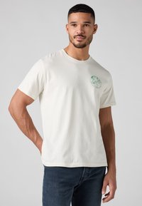 Homme portant un T-shirt blanc avec un globe vert et le logo "Levi's" sur la poitrine gauche, associé à un jean bleu foncé, sur un fond uni.