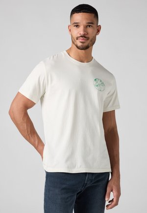 Man in een wit T-shirt met een groene globe en het "Levi's" logo op de linker borst, gecombineerd met donkerblauwe jeans voor een effen achtergrond.