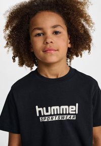 Svart t-shirt i bomull med "hummel SPORTSWEAR"-logotyp i vitt. Korta ärmar, rund halsringning och mjuk textur.