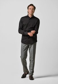 Zwarte lange mouwen shirt, grijze broek en donkere loafers. Het shirt is dichtgeknoopt met een gladde textuur; de broek is op maat gemaakt met een aansluitende snit.