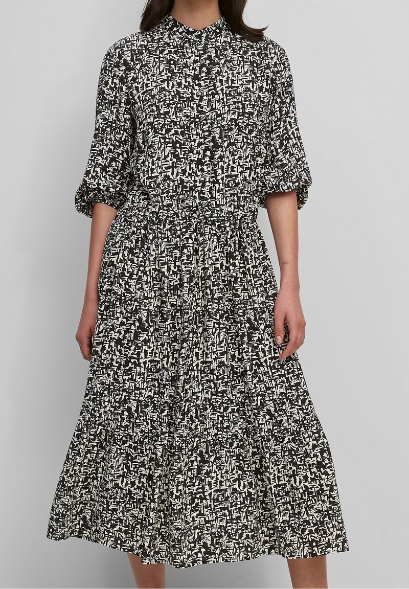 Zwart-witte jurk met patroon, hoge kraag, korte mouwen, getailleerde taille en een vloeiende rok die tot halverwege de kuit reikt.