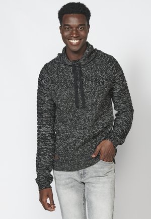 Schwarz-weiße melierte Strickkapuze mit Kordelzugkragen, gerippten Bündchen und strukturiertem Design, kombiniert mit hellgrauen Jeans.