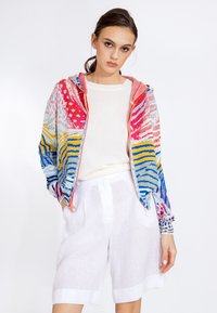 Colorful knit jacket with vivid patterns in pink, blue, and yellow; features a zip front and a lined hood, paired with white linen shorts.
