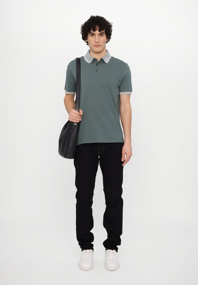 Grønn polo skjorte med grå krage, svart bukser og hvite joggesko. Holder en svart skulderveske. Krøllete hår. Enkel, ren design.