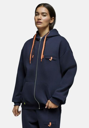 Femme portant un sweat à capuche bleu marine zippé avec des cordons orange et un pantalon bleu marine assorti, debout devant un fond blanc.