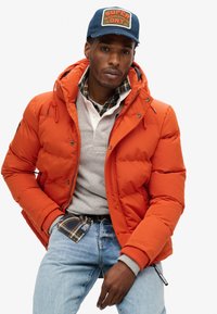 Orange polstret jakke med hætte, grå polo under, ternet skjorte synlig ved kraven, og lyseblå jeans. Baseballkasket med logo.