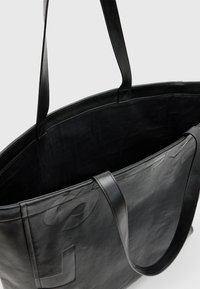 Grand sac fourre-tout en cuir noir avec deux longues poignées, affichant une texture douce et un détail de logo embossé à l'avant. L'intérieur est en tissu noir.