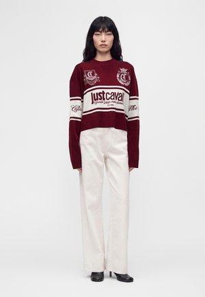 Femme asiatique aux longs cheveux noirs portant un pull Just Cavalli bordeaux et blanc, un pantalon blanc à jambes larges et des chaussures noires à talons, debout.