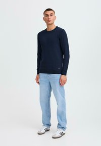 Marineblauer Strickpullover mit Rundhalsausschnitt und gerippten Bündchen, kombiniert mit hellblauen Jeans und weißen Sneakers mit schwarzen Streifen.