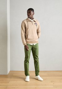 Beige zip-up hoodie featuring an embroidered design, paired with green slim-fit pants and white sneakers. Simple, casual outfit.