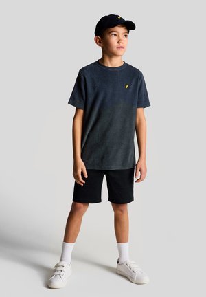 Zēns valkā melnu cepuri, tumši pelēku T-kreklu ar dzeltenu logotipu, melnas šortus, baltas zeķes un baltas sporta kurpes, stāv un skatās pa labi.