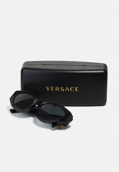 Mustad mustad Versace prillid, millel on nurgelised raamid ja tumedat värvi läätsed, asetatud elegantse musta karbi kõrvale, millel on kuldne kiri.