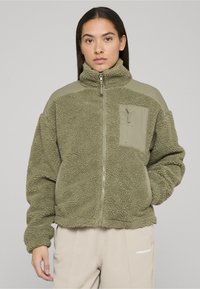 Olīvu zaļa flīsa jaka ar teksturētu virsmu, taisnu apkakli, rāvējslēdzēja aizdari un vienkrāsainu gaiši zaļu kabatu uz kreisās krūtīm.