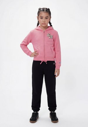 Niña joven con el cabello trenzado, vestida con una sudadera rosa con cremallera y pantalones deportivos negros, de pie con una mano en la cadera.