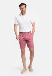 Roze katoenen shorts met zijzakken, gecombineerd met een aansluitend wit poloshirt. Model draagt beige instappers. Effen achtergrond.