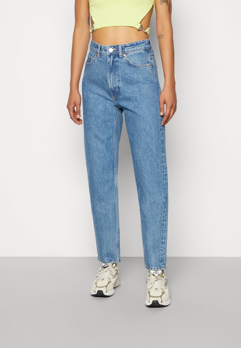 Personne portant un jean droit taille haute bleu clair, un crop top jaune pâle et des baskets blanches Adidas, debout devant un arrière-plan uni.