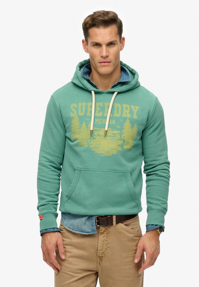 Sudadera verde con un bolsillo canguro en el frente, capucha con cordones y un diseño gráfico amarillo que presenta árboles y agua. Lleva puesta sobre una camisa de mezclilla.