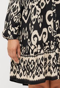 Gros plan sur une main sur une robe à motifs présentant des couleurs noir et crème avec des designs complexes, mettant en valeur un tissu texturé et un ourlet volanté.
